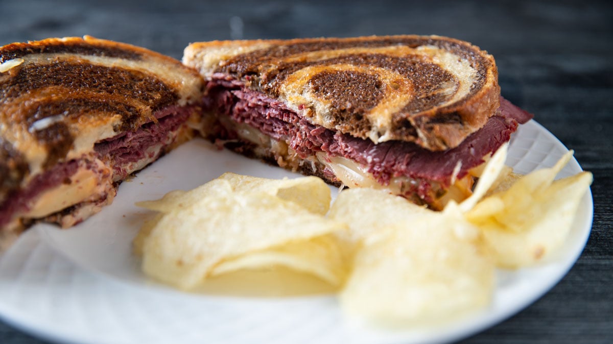 Meaty venison Reuben on a white plate with potato chips.
