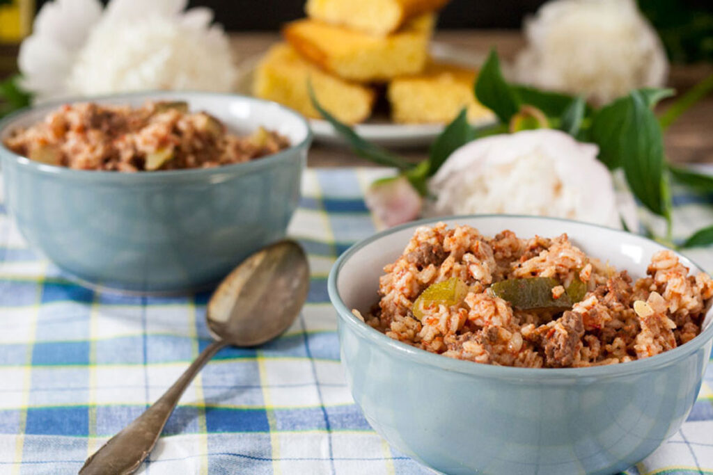 Two blue ceramic bowls filled with a hearty rice and meat dish, possibly jambalaya, are placed on a checkered tablecloth. A spoon rests beside one bowl. In the background, pieces of cornbread and white flowers are partially visible.