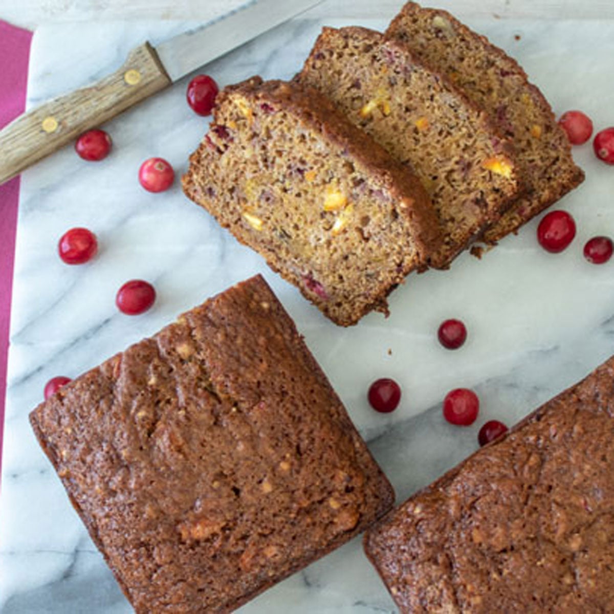 Sliced orange cranberry, pumpkin quick bread on a marble board with strewn cranberries.