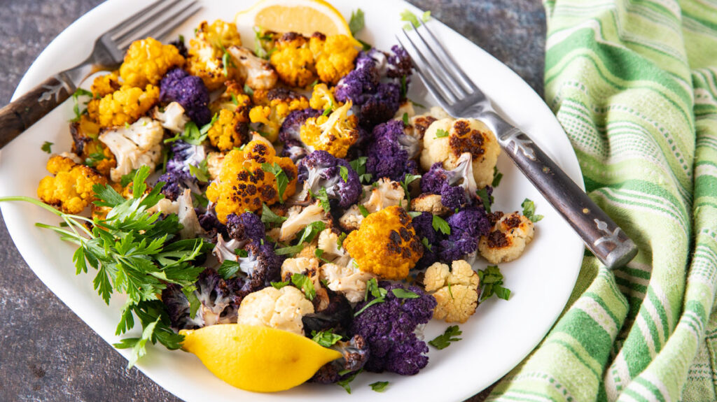 A white plate filled with roasted colorful cauliflower florets in purple, yellow, and white, garnished with fresh parsley and lemon wedges. Two forks are placed on the plate, and a green striped cloth is beside it.
