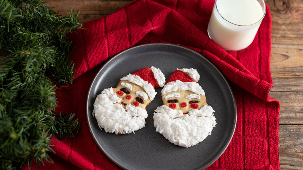 A plate with two Santa-shaped cookies featuring red hats and white beards, alongside a glass of milk. The setup is on a red cloth with green festive foliage on a wooden table.