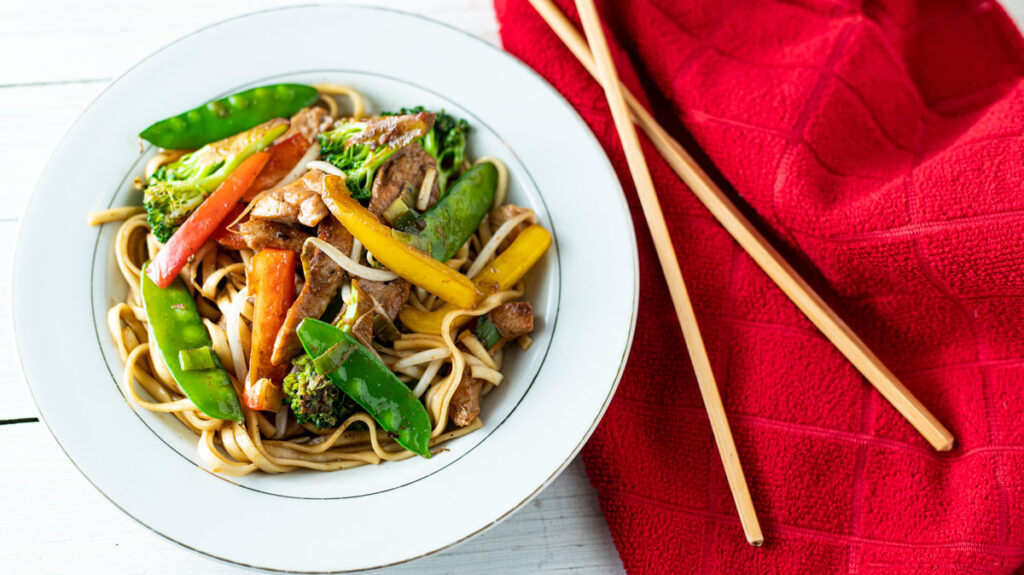 A plate of lo mein noodles topped with stir-fried vegetables including snap peas, bell peppers, broccoli, and strips of meat. The dish is served on a white plate, placed on a white wooden surface next to a pair of wooden chopsticks and red cloth napkin.