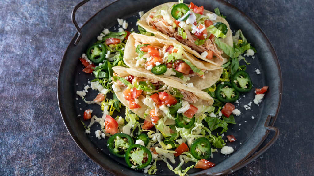 A black plate holds three soft-shell tacos filled with shredded chicken, diced tomatoes, sliced jalapeños, and shredded lettuce. The tacos are topped with crumbled white cheese and garnished with additional sliced jalapeños and lettuce around the edges.