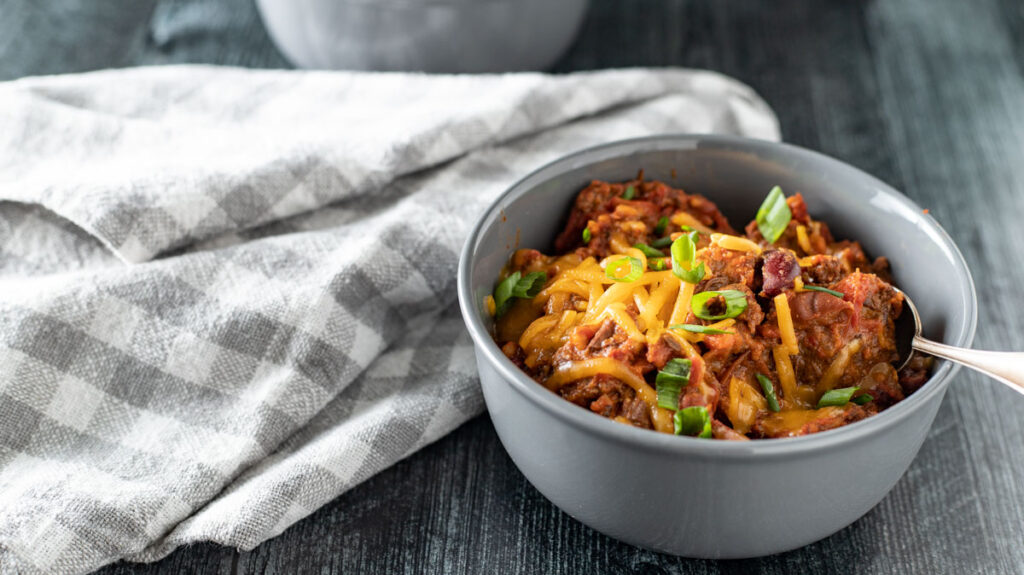 A gray bowl filled with a hearty chili, topped with shredded cheese and chopped green onions. A spoon is placed inside the bowl. The bowl is on a dark wooden table, next to a folded white and gray checkered cloth. Another dish is partially visible in the background.