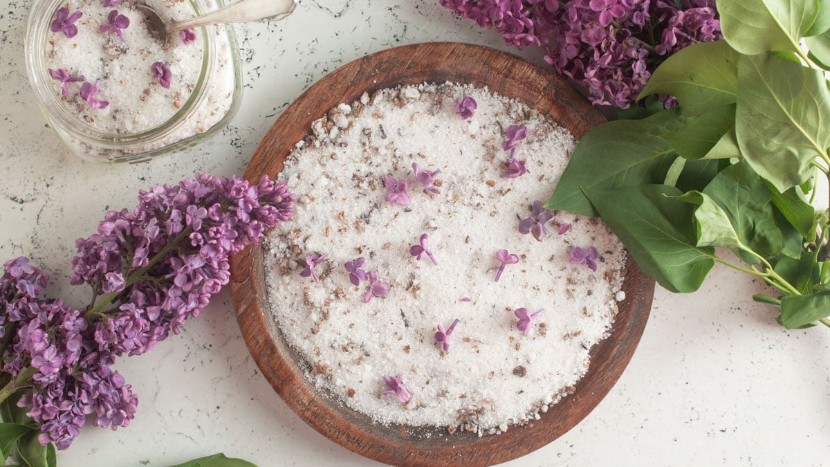 Lilac jelly on a round wooden plate surrounded by lilac flowers and leaves.