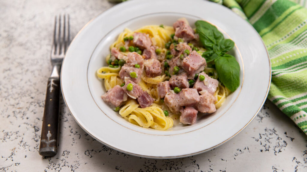 A plate of fettuccine pasta with diced ham and green peas garnished with a basil leaf. The dish is placed on a white textured background, with a fork to the side and a green striped cloth napkin nearby.