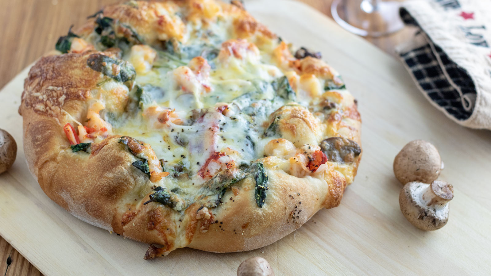 A freshly baked pizza topped with melted cheese, spinach, mushrooms, and diced tomatoes sits on a wooden cutting board. Surrounding the pizza are some uncooked mushrooms and a checkered kitchen towel in the background.