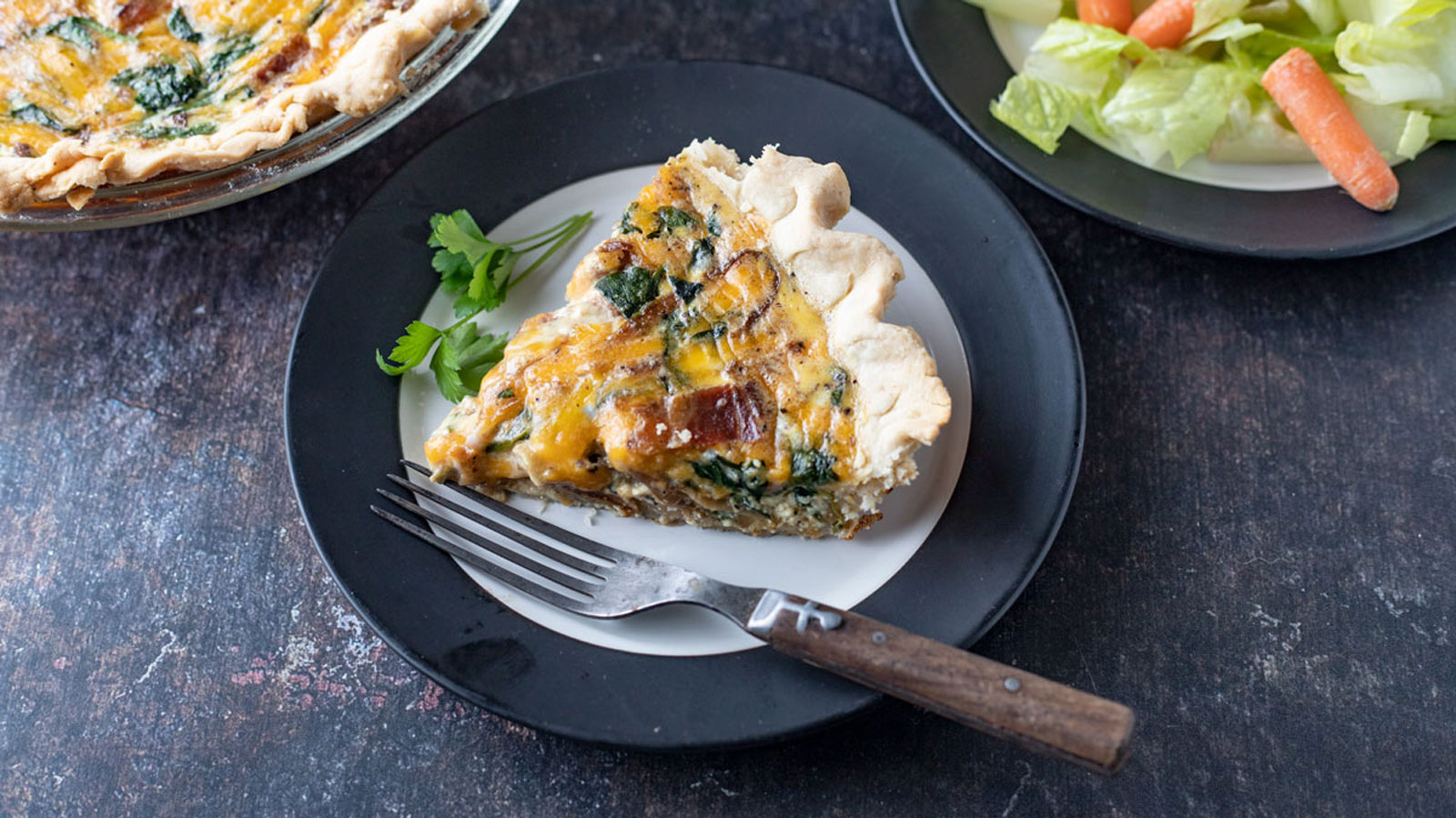 A slice of vegetable quiche with a golden-brown crust and mixed greens on a white plate with a fork. The plate sits on a black dish. Next to it is a salad of romaine lettuce and baby carrots on a grayish background.