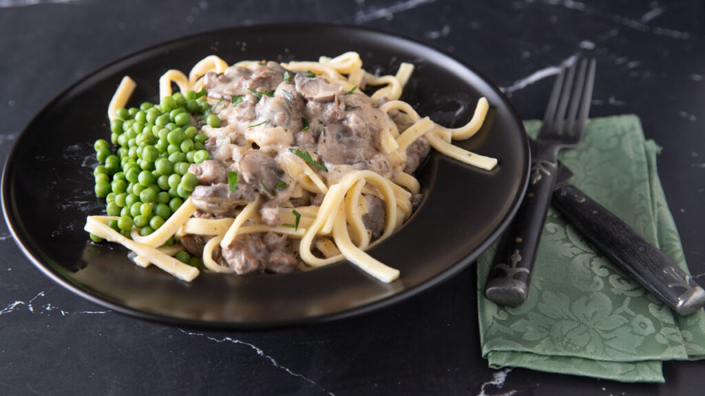 A black plate contains a serving of fettuccine pasta topped with creamy mushroom sauce and a side of green peas. The plate is on a dark marbled surface, accompanied by a fork and knife resting on a folded green napkin with an intricate pattern.