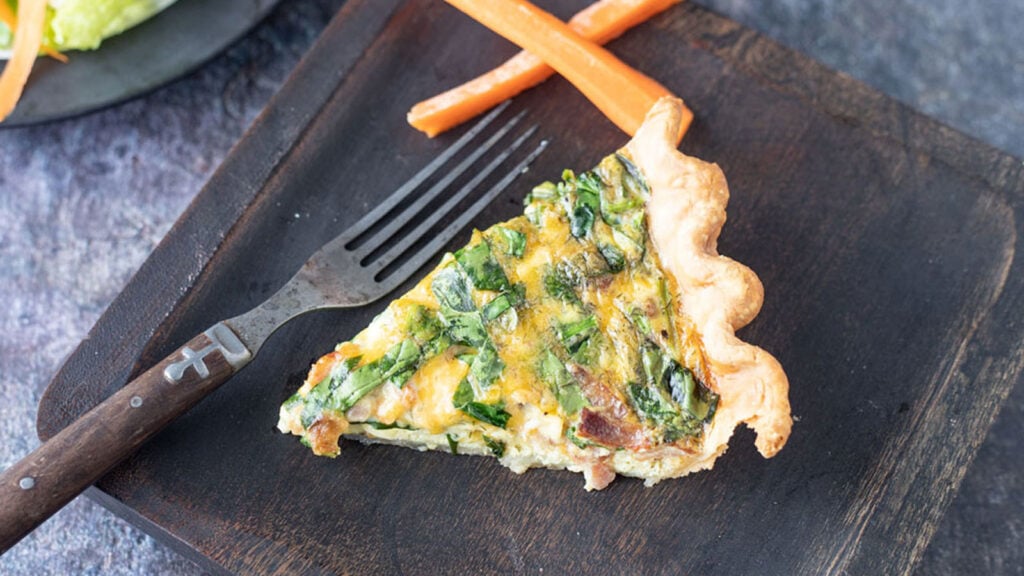 Slice of quiche on a wooden plate with carrot sticks.