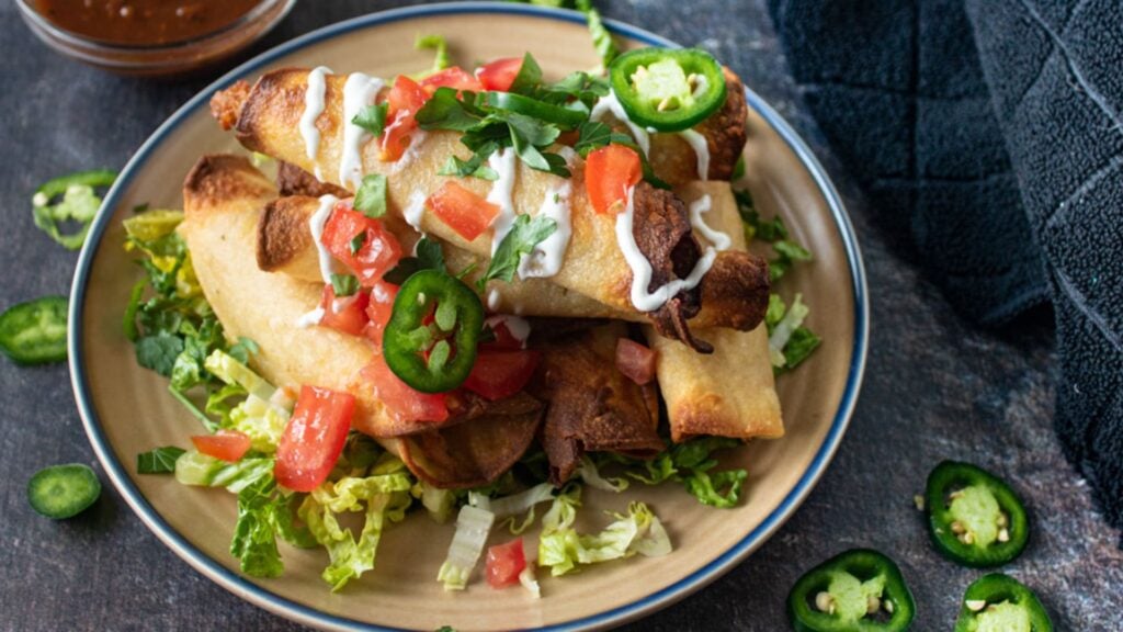 Pile of taquitos garnished with creme and vegetable.
