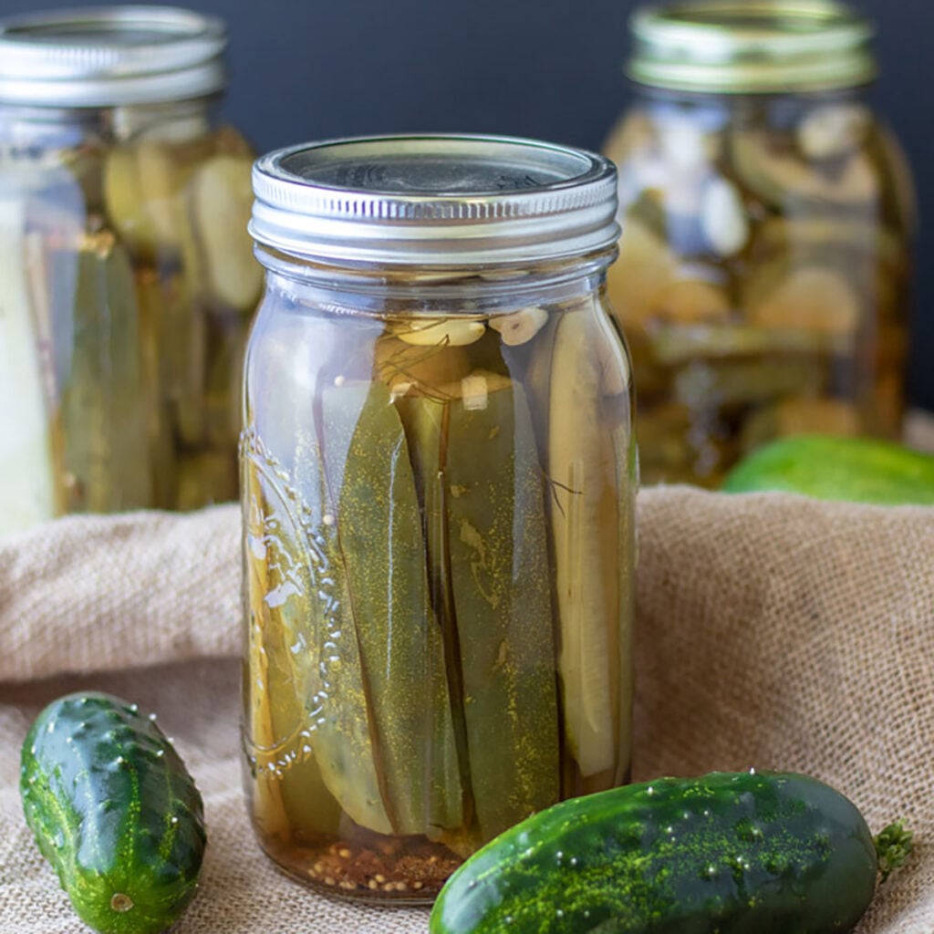 Jars of kosher dill pickles next to fresh cucumbers.