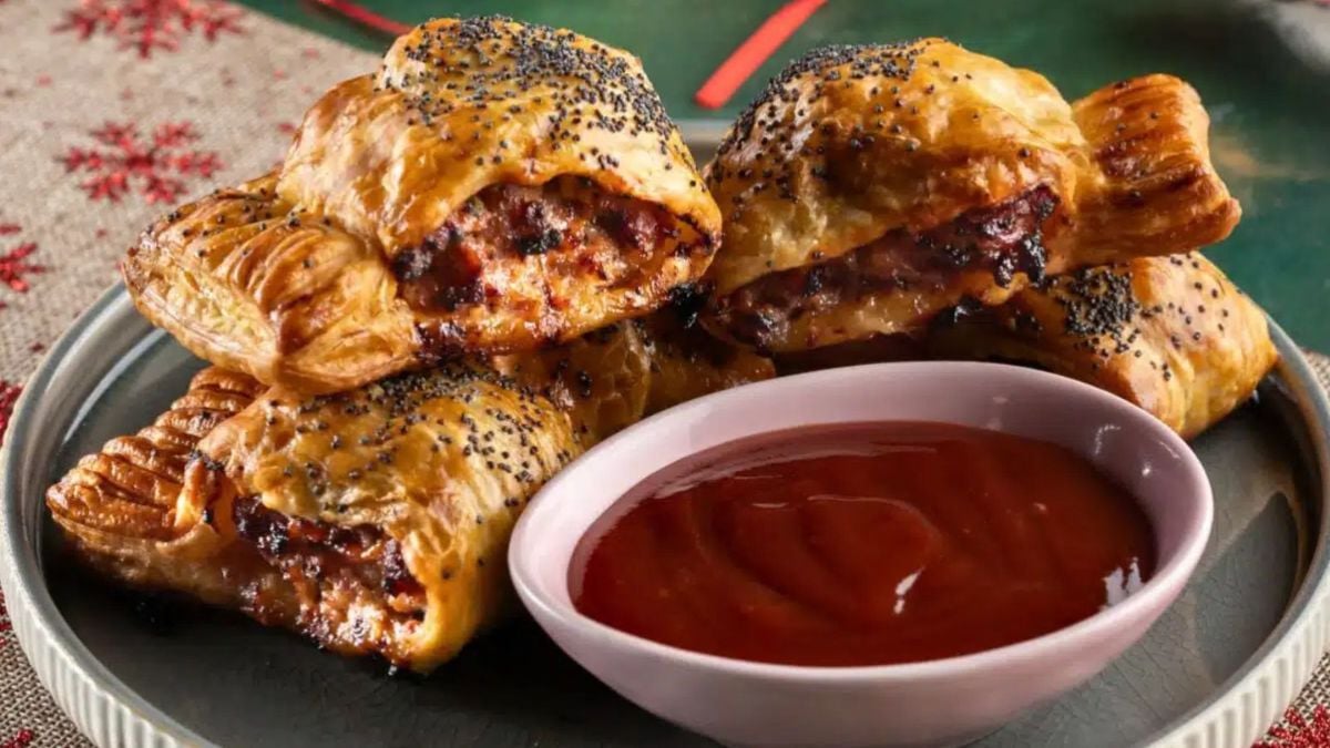 Golden puff pastry apple and sage sausage rolls topped with poppy seeds, served with a bowl of ketchup.