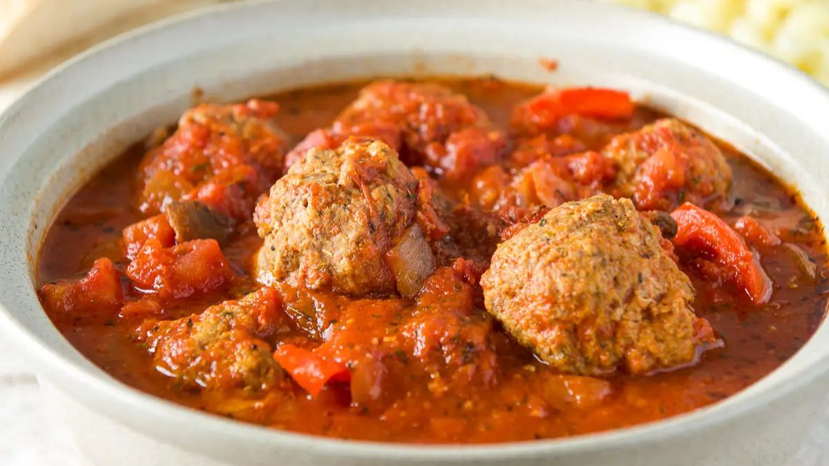 A grey bowl with sauce and meatballs with pasta and bread behind