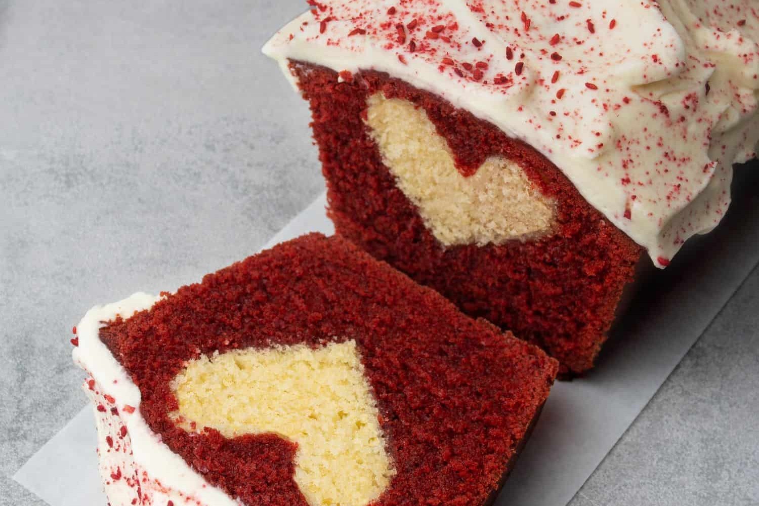 A loaf of Red Velvet Pound Cake with cream frosting on top, sliced to show a surprise heart inside.