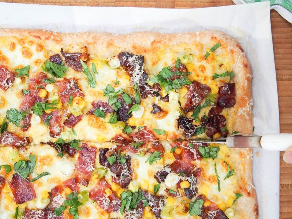 A Bacon Corn Pizza with a knife slicing through it.