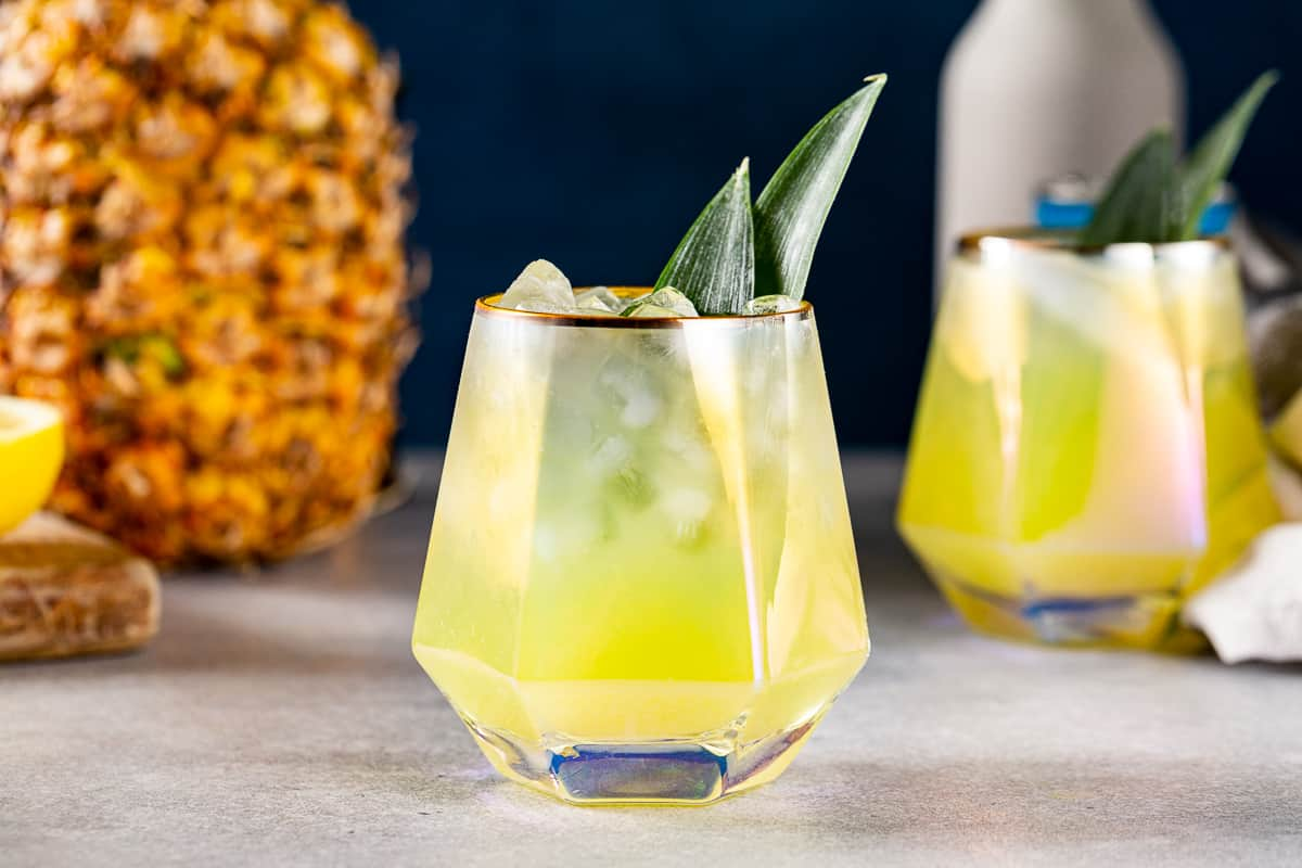 A glass of Pineapple Mocktail with another glass and a pineapple in the background.