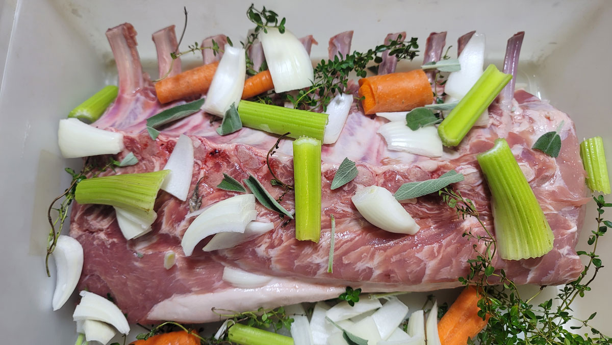 Pork roast in a container with onion, celery, carrots and herbs.