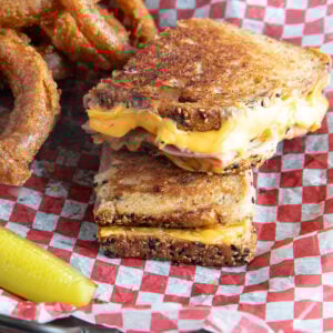 Ham and cheese grilled cheese in a basket with checked paper and a pickle.