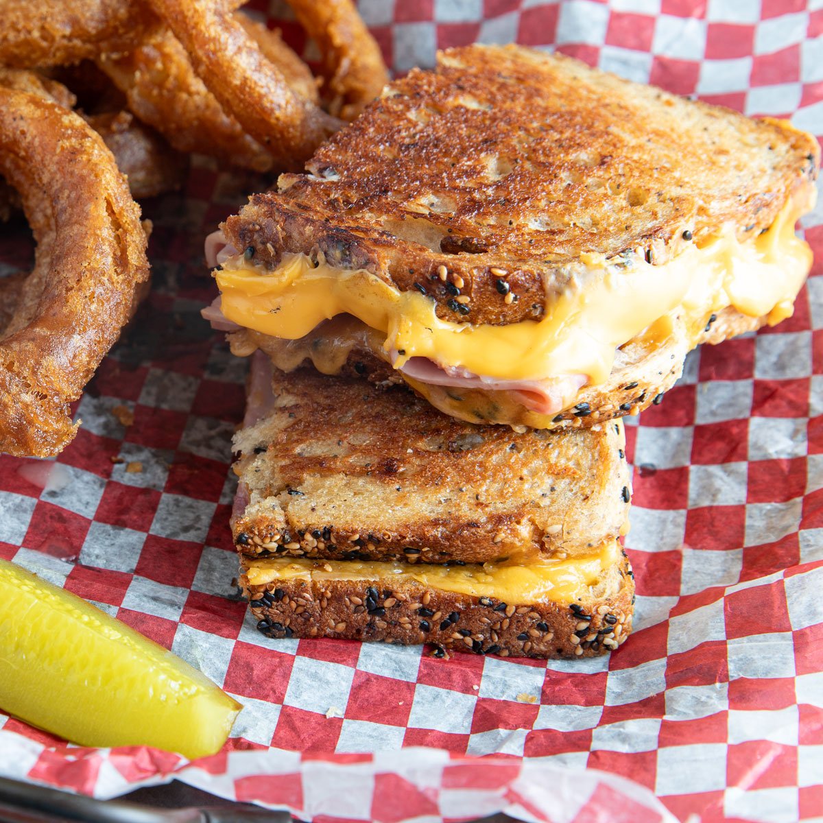 Ham and cheese grilled cheese in a basket with checked paper and a pickle.