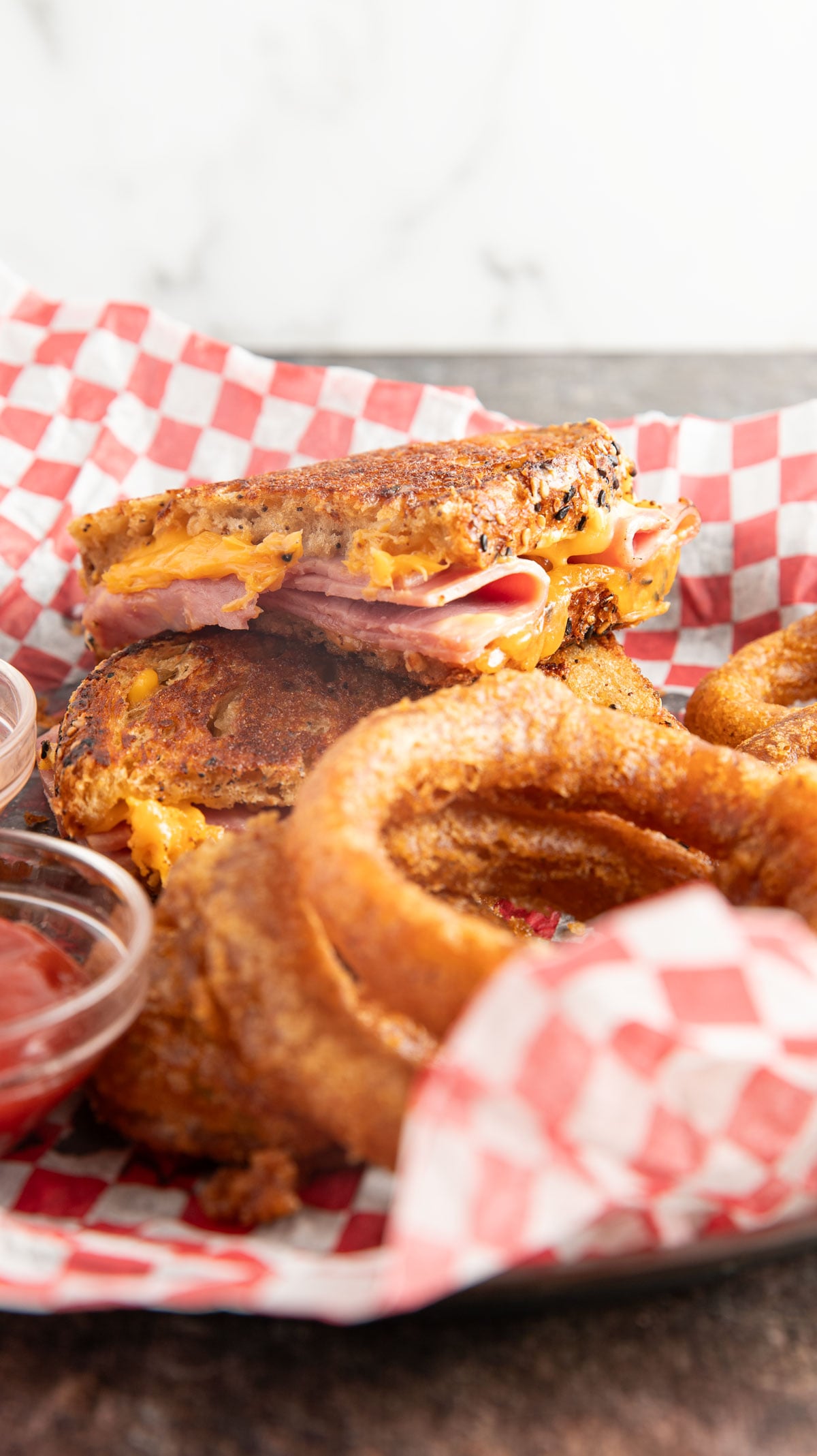 Ham and cheese sandwich in a basket with onion rings of red checked paper.