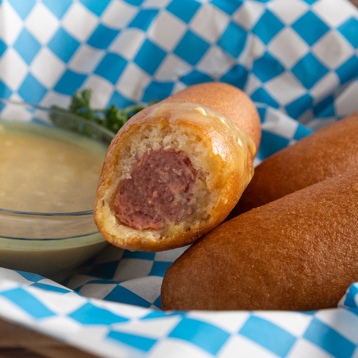 Bite taken out of a corndog in a basket with blue and white checked paper.