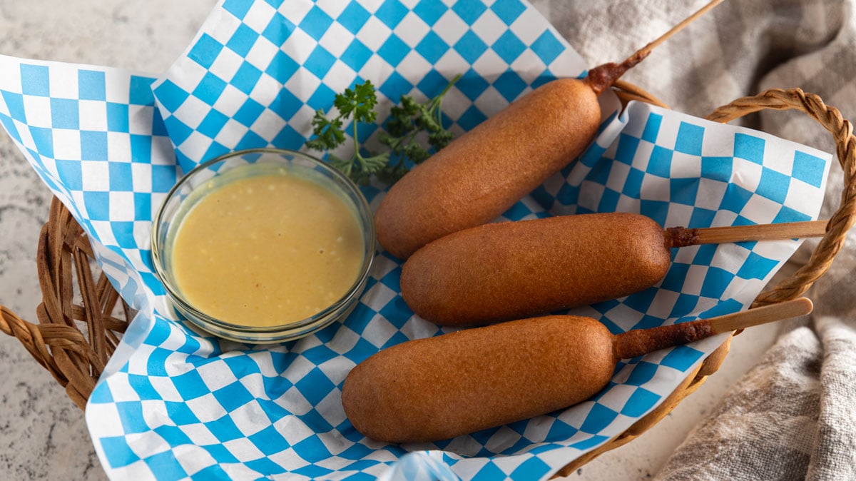 Three air fryer corn dogs in a basket with blue and white checked paper.
