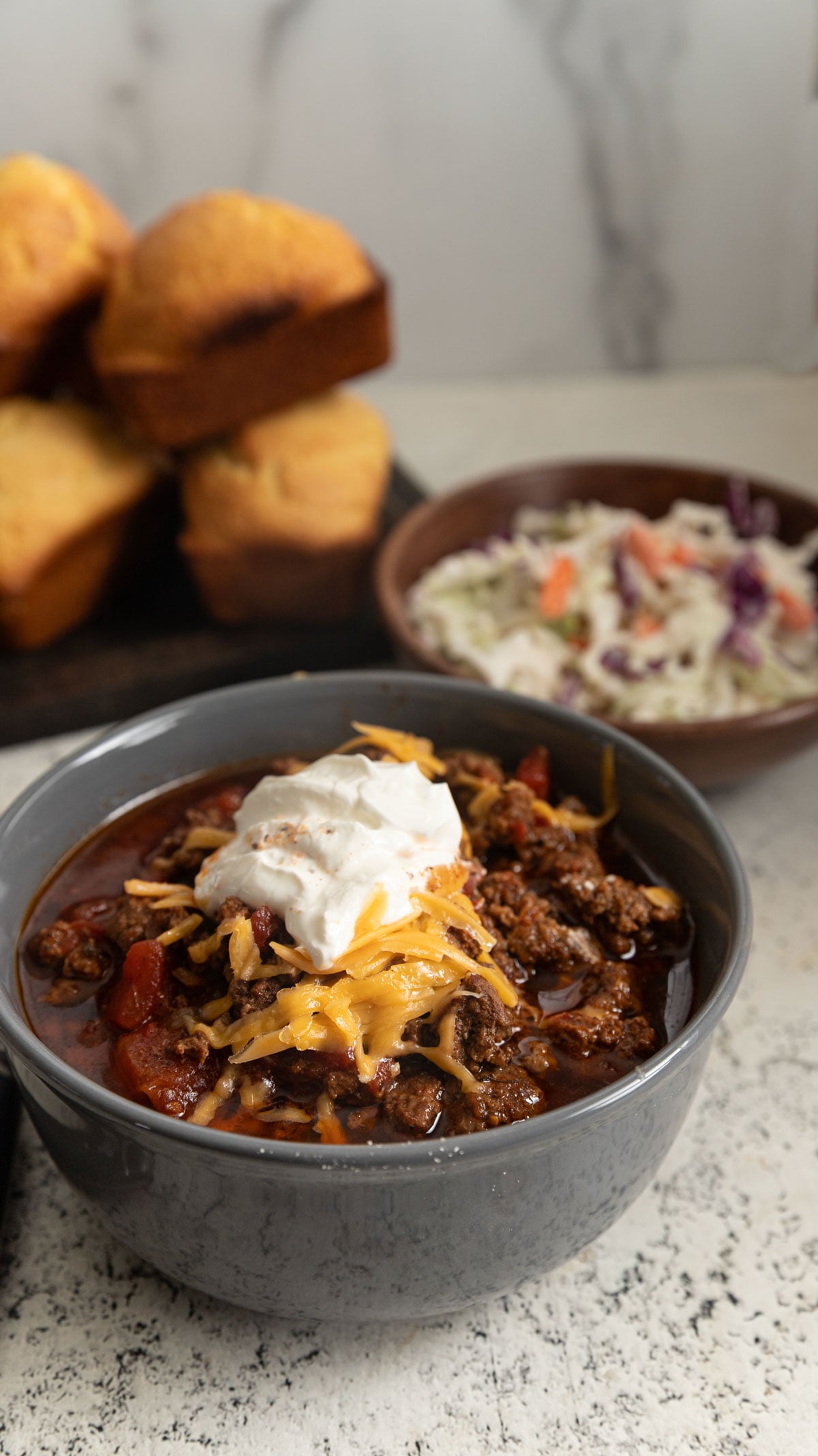 Gray bowl filled with meaty chili topped with grated cheese and sour cream.
