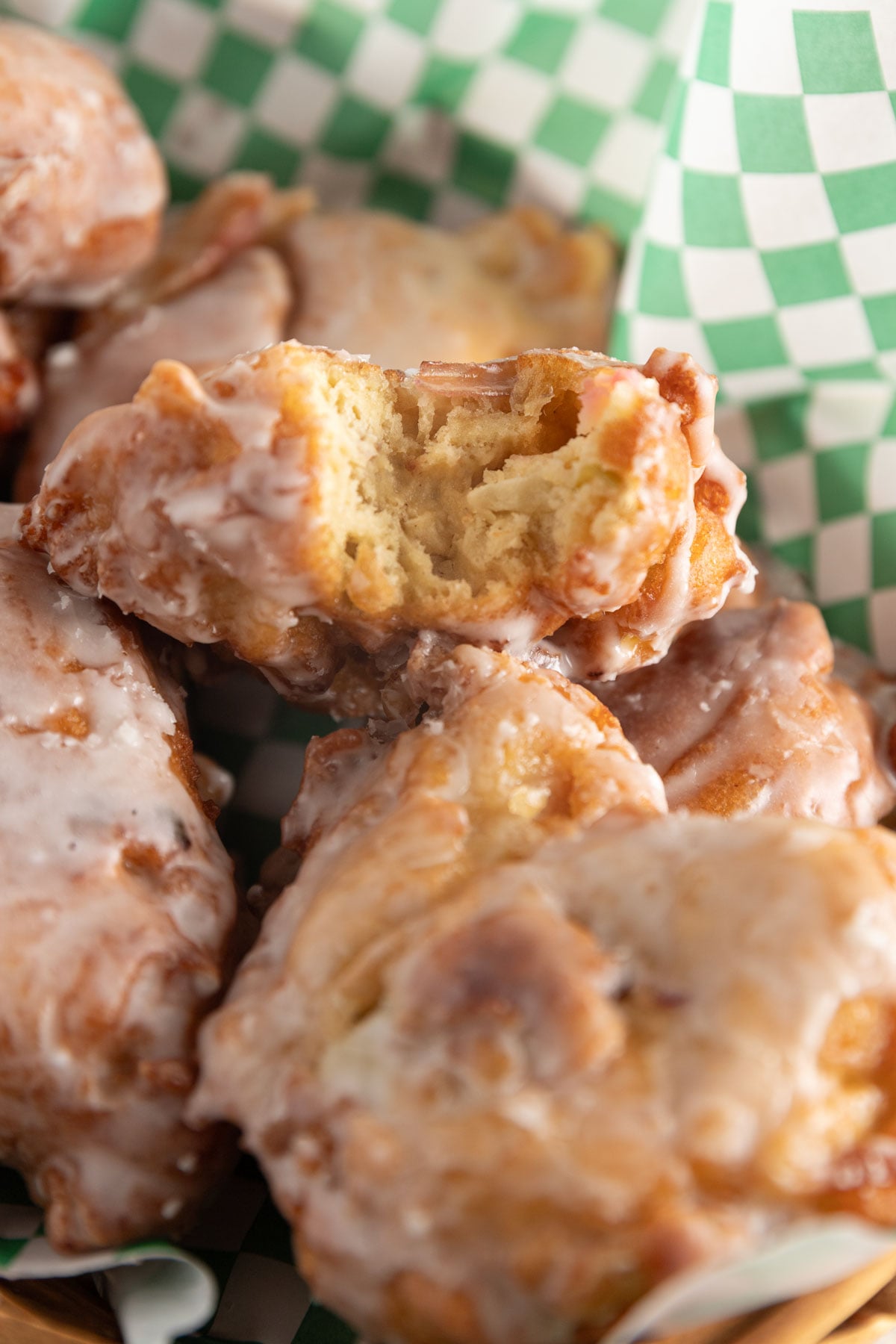 Bite taken out of a an apple fritter in a basket showing the fluffy texture with green and white checked paper.