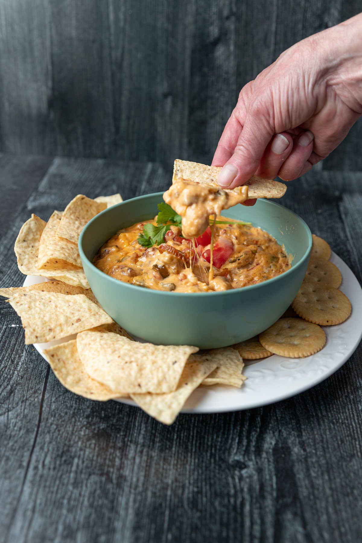Smoked queso dip in a green bowl with a woman's hand scooping a tortilla chips.