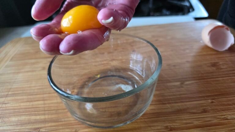 Woman's hand with an egg yolk on her fingers and the egg white draining into a bowl.